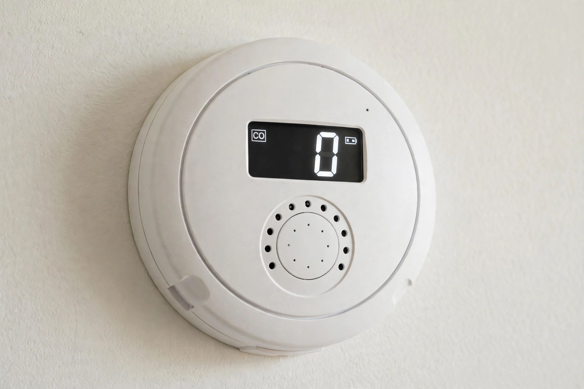 Close-up photograph of a round white carbon monoxide alarm with a digital display, mounted on a light-colored wall