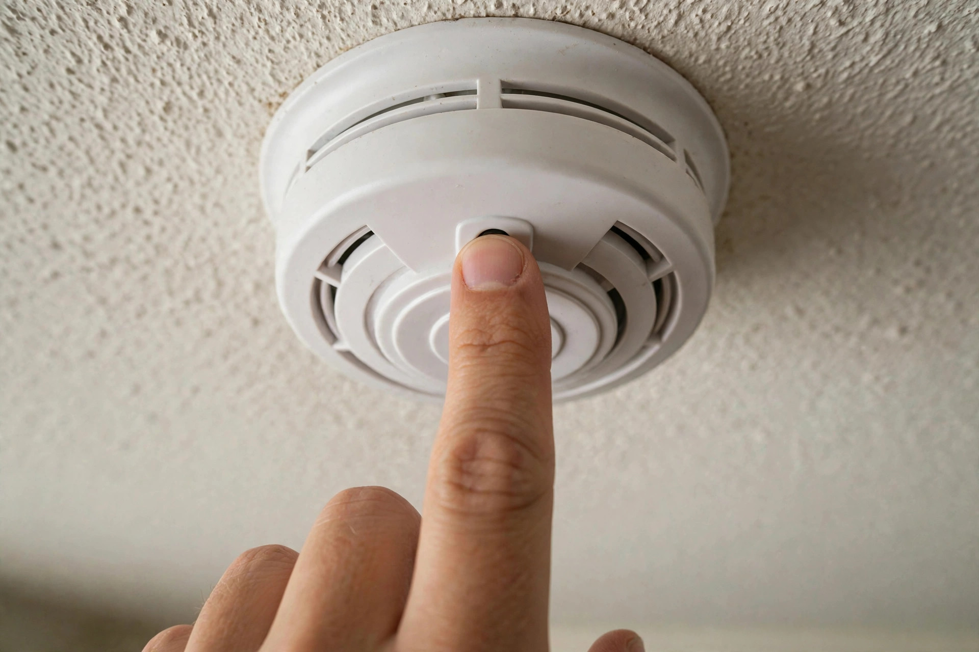 Close-up of fingers pressing the test button on a smoke detector mounted on a ceiling, indoor home setting