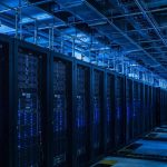 interior of a large data center with rows of server racks and small sampling pipes running along the ceiling, cool blue light