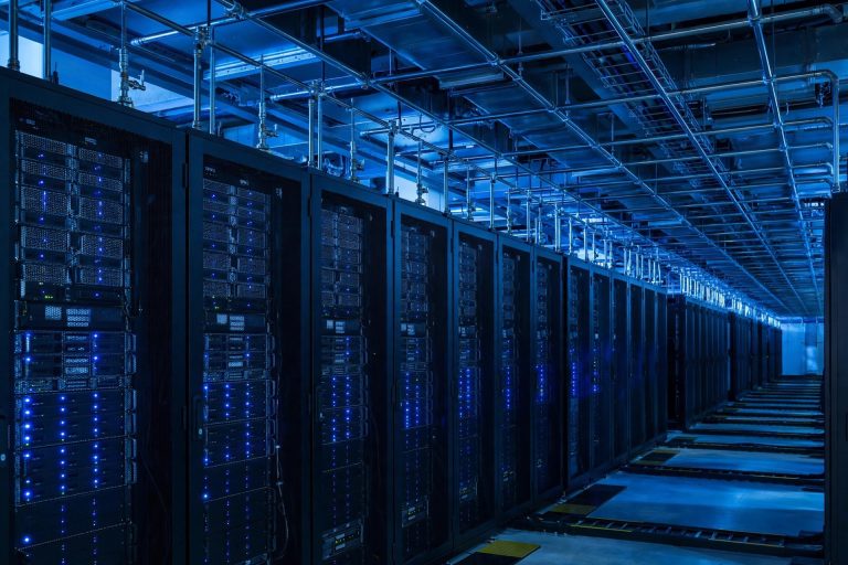 interior of a large data center with rows of server racks and small sampling pipes running along the ceiling, cool blue light
