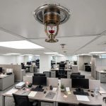 office ceiling with a fire sprinkler head in the foreground and desks below, bright interior lighting